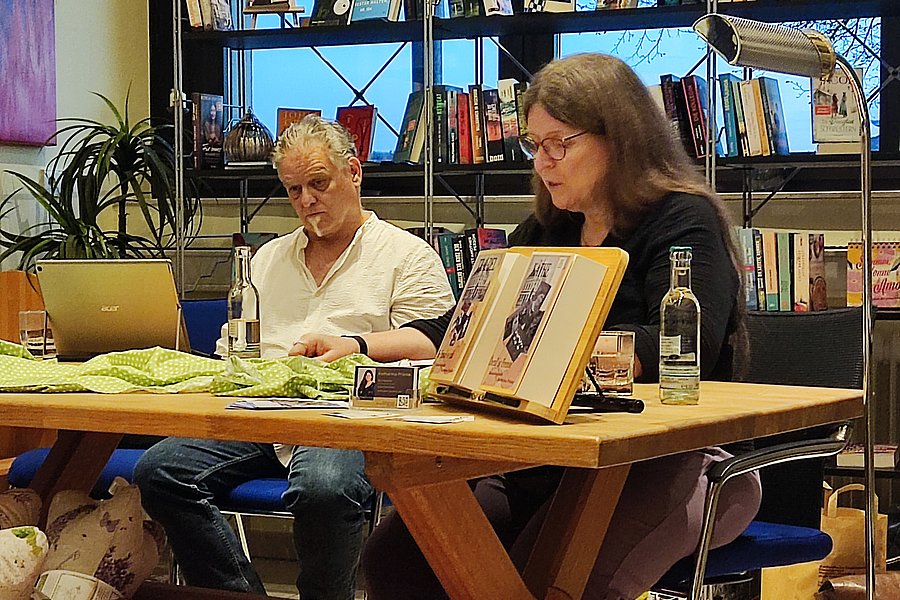 Zwei Personen sitzen an einem Holztisch in einem Raum mit Bücherregalen und lesen beziehungsweise arbeiten