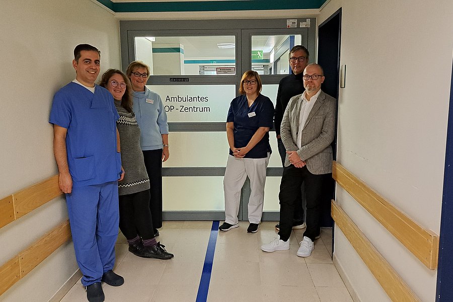 Sechs Personen, darunter medizinisches Personal, stehen in einem Krankenhausflur vor einer Tür mit der Aufschrift 'Ambulantes OP-Zentrum'