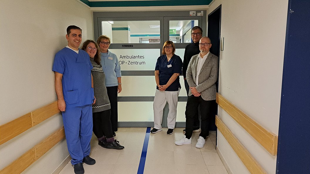 Sechs Personen, darunter medizinisches Personal, stehen in einem Krankenhausflur vor einer Tür mit der Aufschrift 'Ambulantes OP-Zentrum'