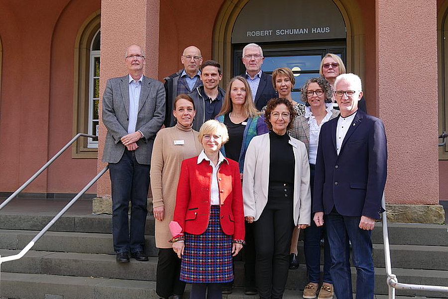 Elf Personen stehen auf Treppen vor dem Gebäude mit der Aufschrift 'Robert Schuman Haus', formell gekleidet, Gruppenfoto bei Tageslicht