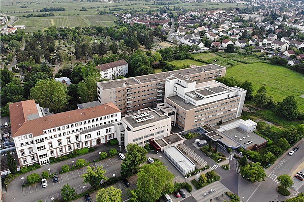 Luftaufnahme eines großen Gebäudekomplexes mit mehreren verbundenen Gebäuden, umgeben von Parkplätzen, Bäumen und einer Wohnsiedlung im Hintergrund