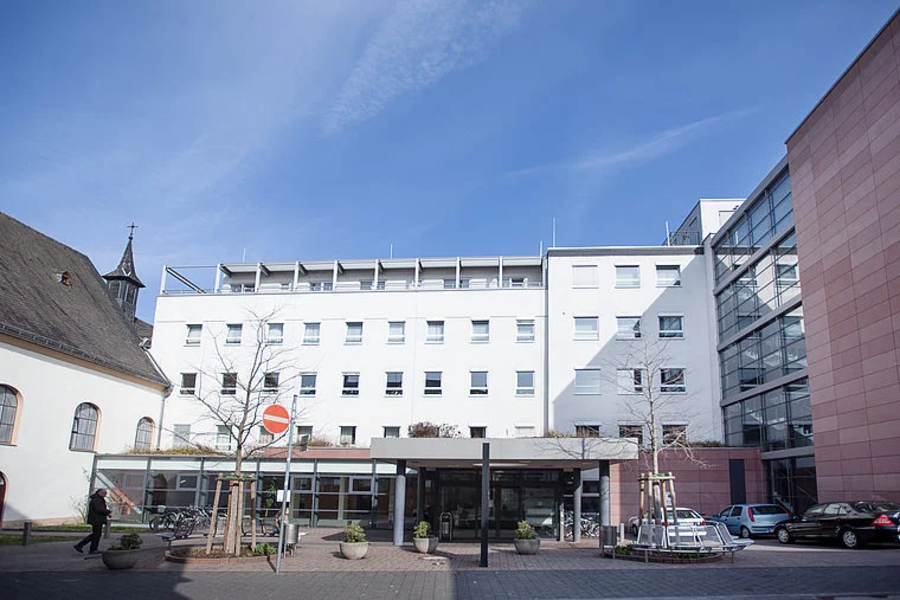 Weißes mehrstöckiges Gebäude mit Glasfassade rechts, davor gepflasterter Platz mit Bäumen und Autos, blauer Himmel