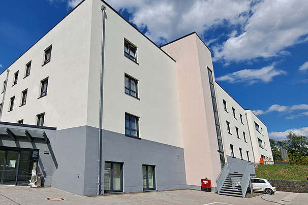 Vierstöckiges modernes Gebäude mit weißen Fassaden, mehreren Fenstern und einem überdachten Eingang bei blauem Himmel.