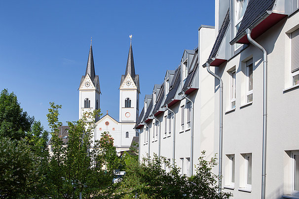 Zwei weiße Kirchtürme mit spitzen Dächern hinter einem weißen Wohngebäude mit roten Dachfenstern und grünen Bäumen im Vordergrund.
