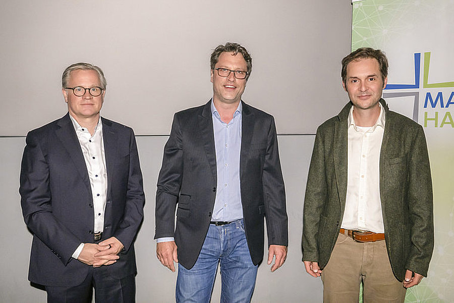 Drei Männer stehen nebeneinander vor einer Wand, zwei tragen Anzüge, einer trägt Sakko und Hemd, alle schauen in die Kamera