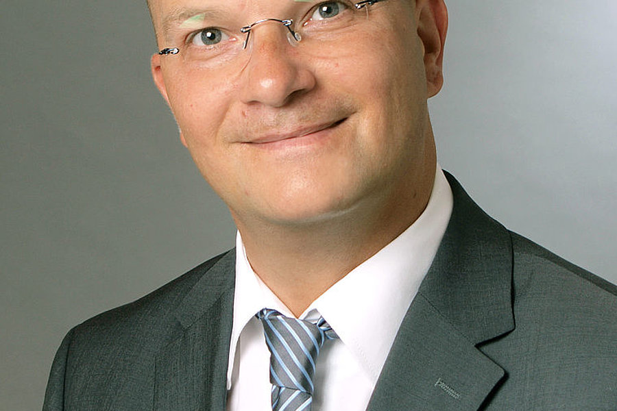 Porträt eines lächelnden Sascha Koenigshoven mit Brille, grauem Anzug, weißem Hemd und gestreifter Krawatte vor neutralem Hintergrund