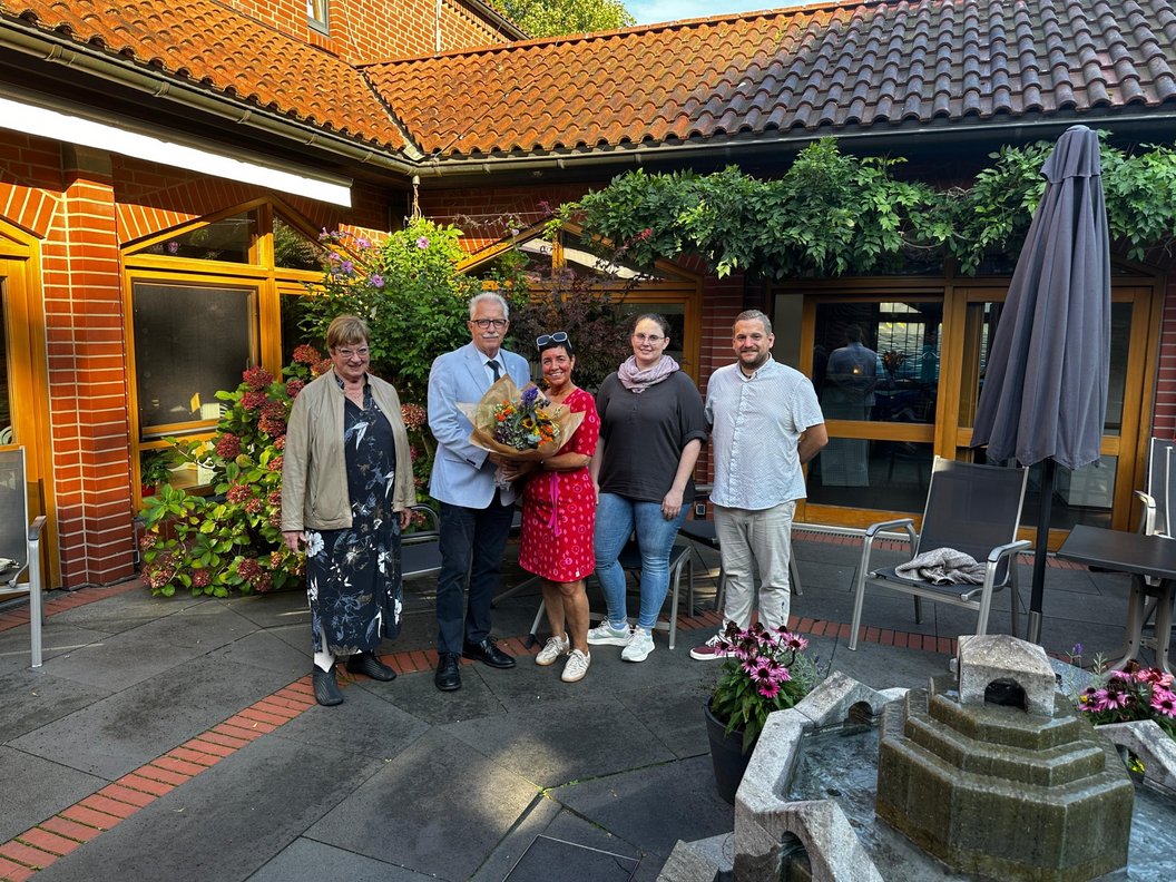 Fünf Personen stehen auf einem gepflasterten Hof vor einem Backsteingebäude, eine Person hält einen Blumenstrauß.