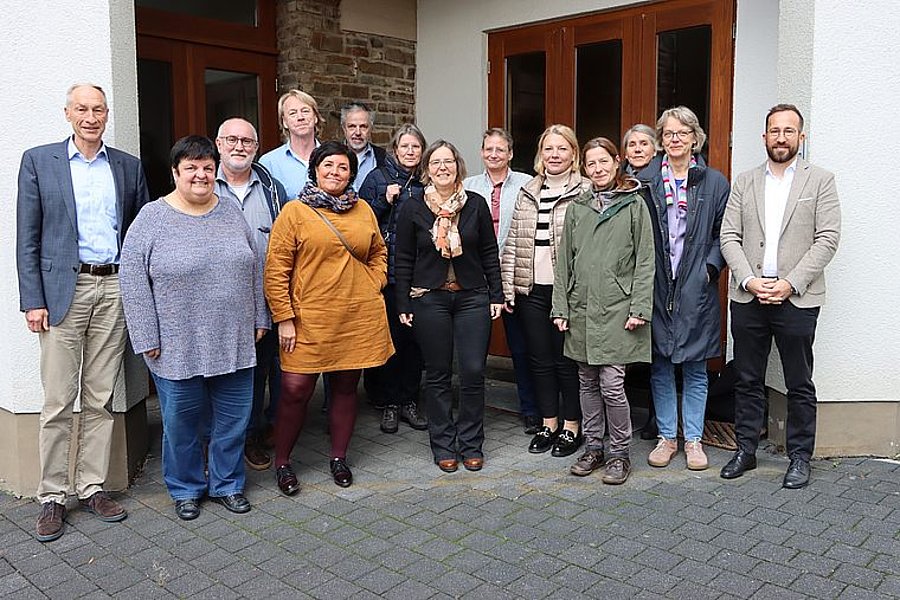 Gruppenfoto von 14 Personen vor einem Gebäude mit weißen Wänden und braunen Türen, alle stehen nebeneinander und schauen in die Kamera