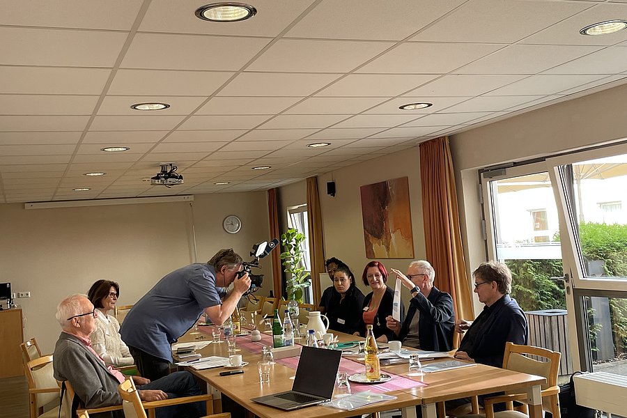 Sechs Personen sitzen an einem Tisch in einem hellen Raum, eine Person fotografiert die Gruppe mit einer Kamera