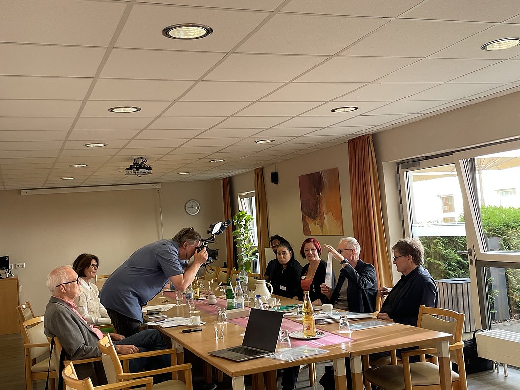 Sechs Personen sitzen an einem Tisch in einem hellen Raum, eine Person fotografiert die Gruppe mit einer Kamera