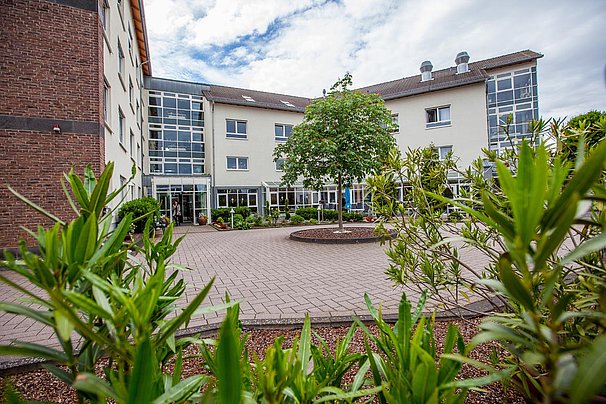 Mehrstöckiges Gebäude mit großen Fenstern und Eingang, umgeben von grünem Pflanzenbeet und gepflastertem Innenhof unter bewölktem Himmel.