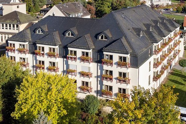 Mehrstöckiges weißes Wohngebäude mit vielen Balkonen, die mit Blumenkästen geschmückt sind, umgeben von Bäumen und Grünflächen.
