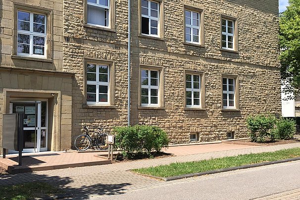 Dreistöckiges Sandsteingebäude in Bitburg mit mehreren Fenstern und einem Eingang links, davor ein Fahrrad und Grünflächen