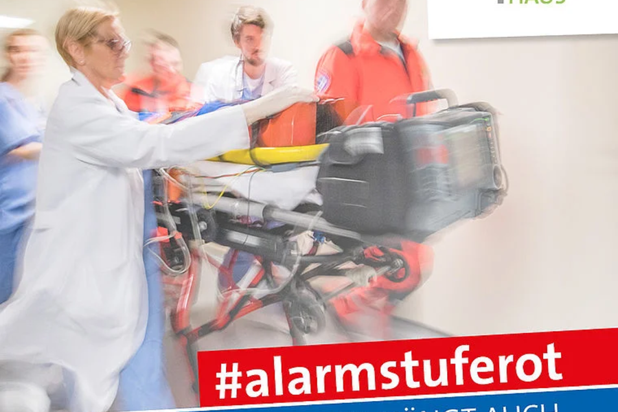 Medizinisches Personal transportiert einen Patienten auf einer Trage im Krankenhaus Marienhaus, Text '#alarmstuferot Im Notfall hängt auch dein Leben davon ab'