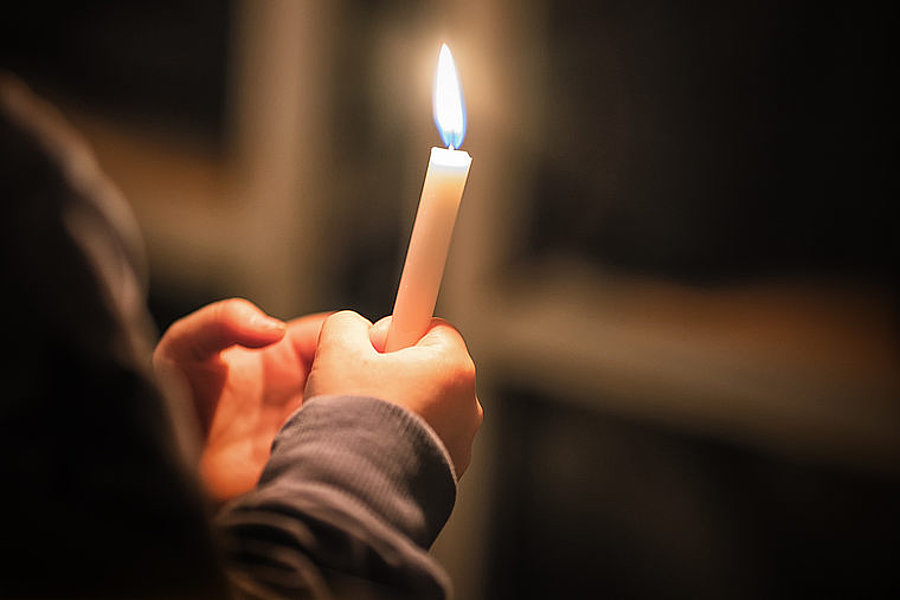 Person hält eine brennende weiße Kerze in der Hand vor dunklem Hintergrund