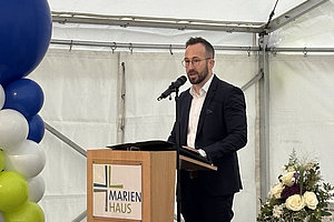 Mann in dunklem Anzug steht hinter Rednerpult mit Mikrofon, links bunte Ballons, rechts Blumengesteck auf weiß gedecktem Tisch