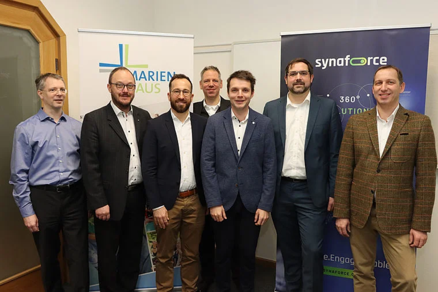 Sieben Männer in formeller Kleidung stehen vor Bannern mit den Logos von Marienhaus und synaforce