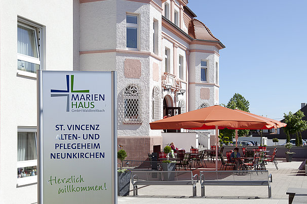 Eingangsbereich eines Pflegeheims mit Schild 'Marienhaus St. Vincenz Alten- und Pflegeheim Neunkirchen Herzlich willkommen!', Sitzbereich mit roten Sonnenschirmen und historisches Gebäude im Hintergrund