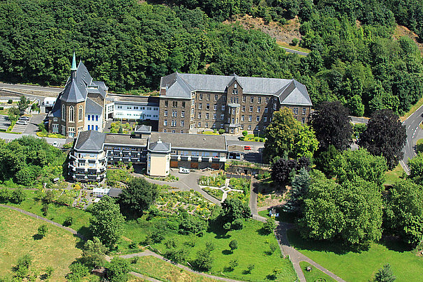Luftaufnahme eines großen historischen Gebäudekomplexes mit Gärten, umgeben von Wald und einer Straße rechts