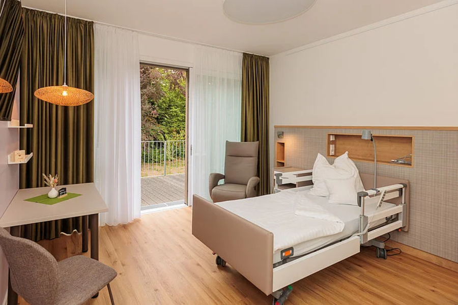 Modernes Patientenzimmer mit Pflegebett, Sessel, Tisch, Stuhl und Zugang zu Balkon mit Blick ins Grüne