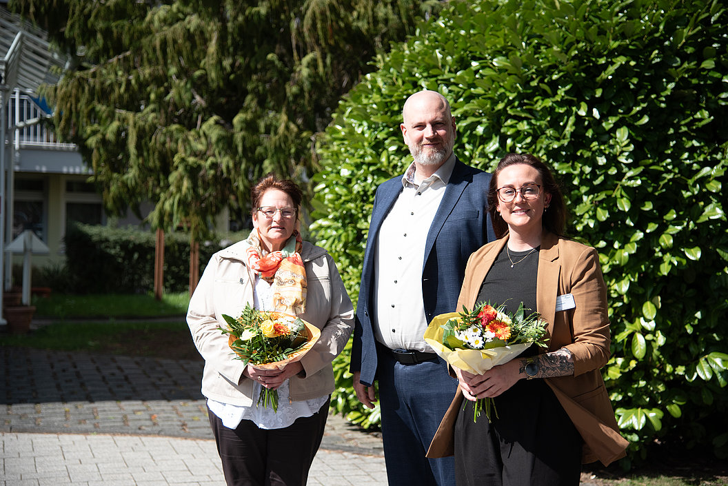 Drei Personen stehen mit Blumensträußen vor einem großen grünen Busch im Freien