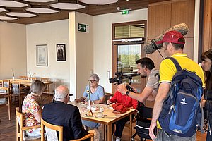 Mehrere Personen sitzen an einem Holztisch in einem Raum mit sechseckiger Deckenverkleidung, während ein Mann mit roter Kappe und blauem Rucksack eine Kamera auf sie richtet.