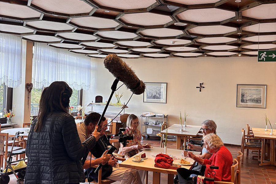 Person mit Mikrofon auf Stab filmt ältere Menschen, die an einem Tisch in einem Raum mit Holzdecke sitzen