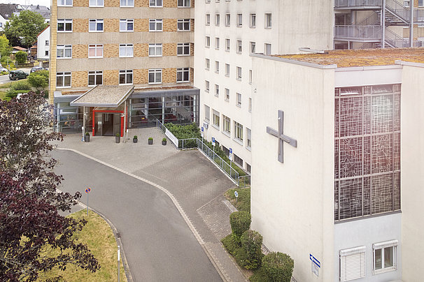 Eingang eines Krankenhausgebäudes mit einem großen Kreuz an der Fassade und mehreren mehrstöckigen Gebäuden im Hintergrund
