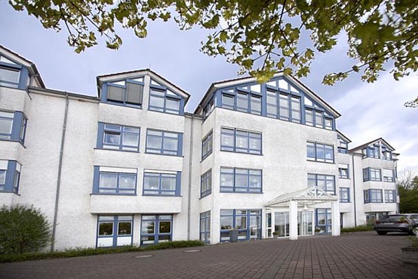 Dreistöckiges weißes Bürogebäude mit blauen Fensterrahmen und Glasvordach am Eingang, umgeben von Bäumen und Parkplatz.