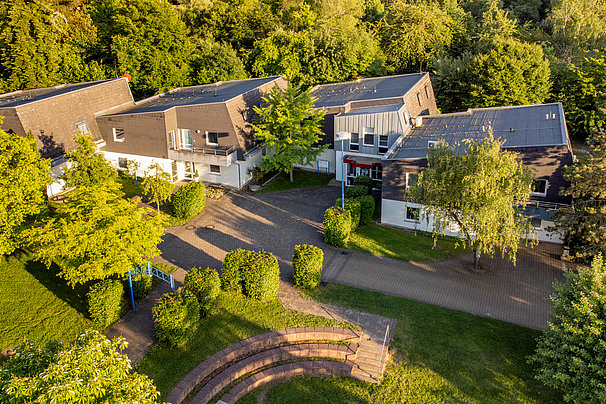Luftaufnahme eines Gebäudekomplexes mit mehreren verbundenen Gebäuden, umgeben von Bäumen und Grünflächen, mit einem halbrunden Amphitheater im Vordergrund