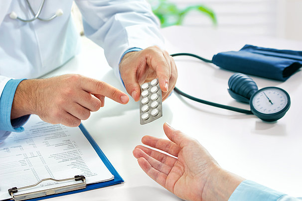 Arzt zeigt Patientin eine Tablettenpackung, auf einem Tisch liegen ein Klemmbrett mit Formular und ein Blutdruckmessgerät.