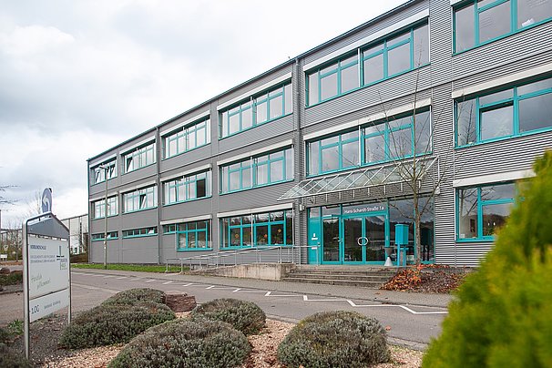 Modernes Bürogebäude mit großen Fenstern und einem Eingang mit Glasvordach, umgeben von bepflanzten Beeten und Parkplätzen
