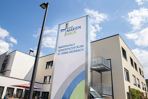 Schild mit dem Schriftzug 'Marienhaus Seniorenhaus Seniorenzentrum St. Anna Morbach' vor einem mehrstöckigen Gebäude mit Balkonen und einem Laternenmast bei blauem Himmel.