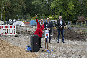 Person in rotem Mantel steht vor Mikrofon und Lautsprecher auf einer Baustelle, zwei weitere Personen stehen im Hintergrund.