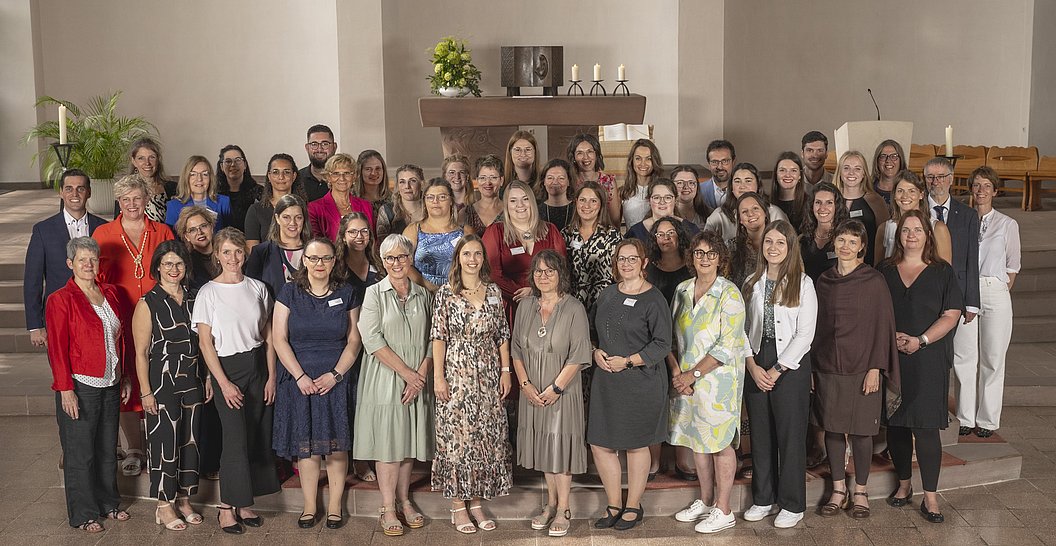 Gruppenfoto von 38 Personen in einem Raum mit beige Wänden, einige tragen formelle Kleidung, andere legere, im Hintergrund ein Kamin mit Dekoration.