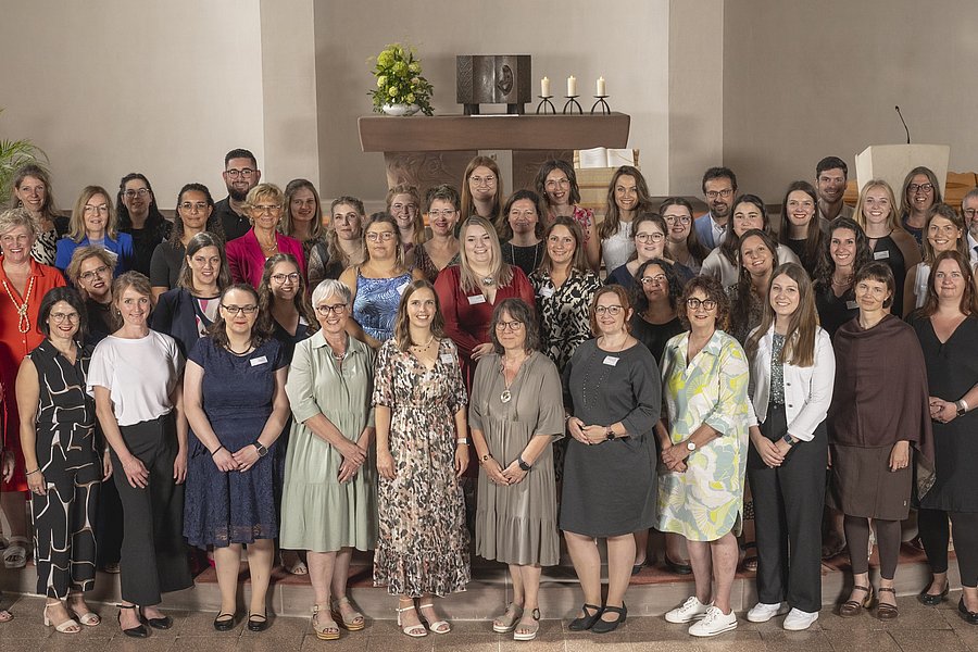 Gruppenfoto von 38 Personen in einem Raum mit beige Wänden, einige tragen formelle Kleidung, andere legere, im Hintergrund ein Kamin mit Dekoration.