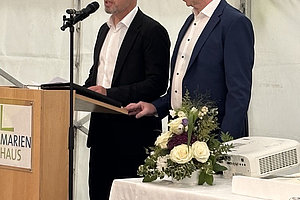 Zwei Männer in Anzügen stehen an einem Rednerpult mit Mikrofon in einem Zelt, daneben ein Tisch mit Blumenstrauß und Projektor.