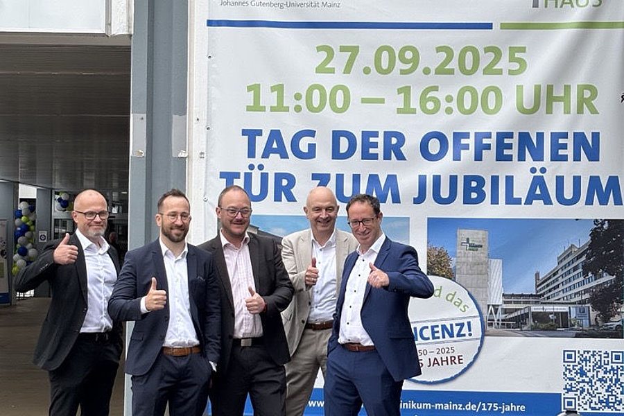 Vier Männer in Anzügen posieren vor einem Plakat mit der Aufschrift '27.09.2025 11:00 – 16:00 UHR TAG DER OFFENEN TÜR ZUM JUBILÄUM' am Marienhaus Klinikum Mainz.