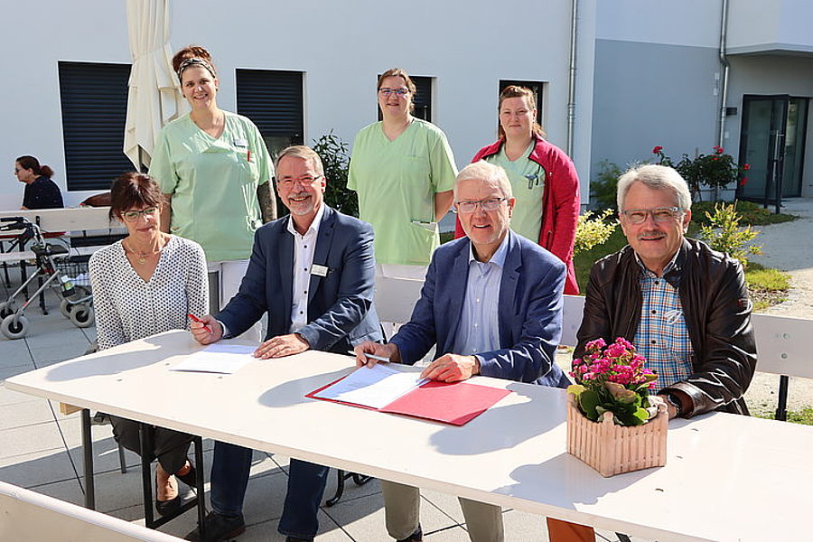 Drei Männer und eine Frau sitzen an einem weißen Tisch im Freien, unterschreiben Dokumente, drei Frauen in medizinischer Kleidung stehen dahinter