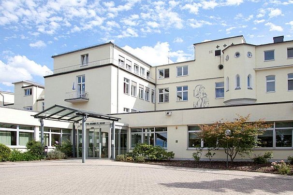 Eingangsbereich eines weißen mehrstöckigen Gebäudes mit Glasvordach und Büschen vor dem Eingang unter blauem Himmel