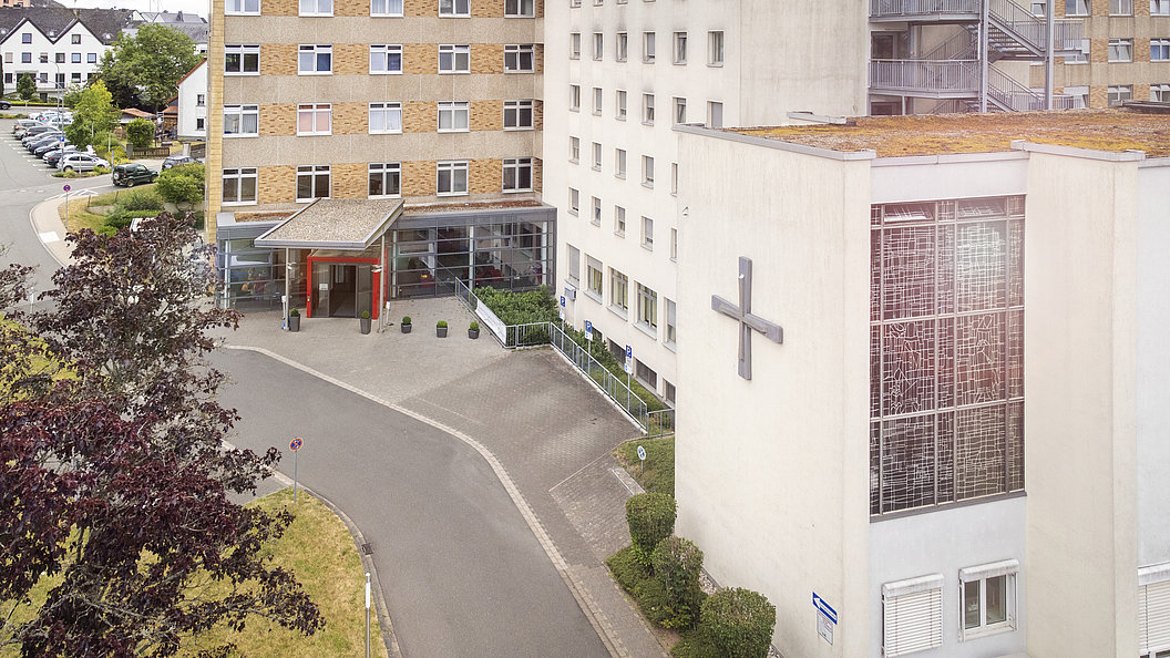 Außenansicht eines mehrstöckigen Gebäudekomplexes mit einem großen Kreuz an der Fassade und einem überdachten Eingang