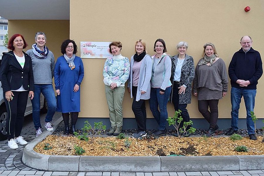 Neun Personen stehen vor einer gelben Wand mit einem Schild 'Hospiz SAPV', sie tragen lässige Kleidung und lächeln