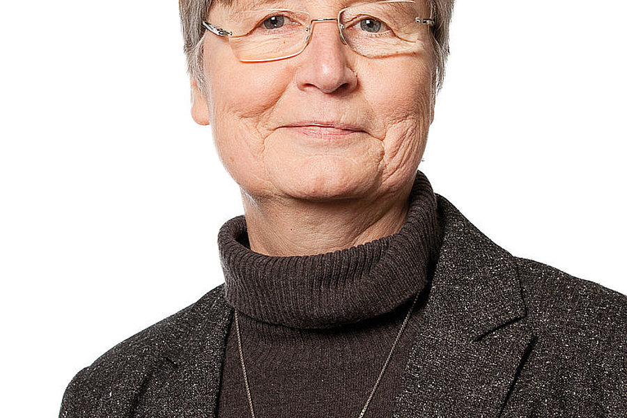 Porträt von Schwester Scholastika, ältere Frau mit kurzen grauen Haaren, Brille, dunklem Rollkragenpullover und Kreuzkette