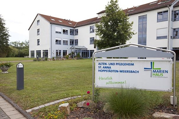 Gebäude eines Alten- und Pflegeheims mit mehrstöckigem Bau, Rasenfläche und Schild mit der Aufschrift 'Alten- und Pflegeheim St. Anna Hoppstädten-Weiersbach Marienhaus Herzlich willkommen'