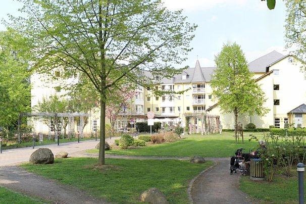 Park mit Wegen, Bänken und Bäumen vor einem mehrstöckigen Gebäude mit spitzem Dach im Hintergrund.