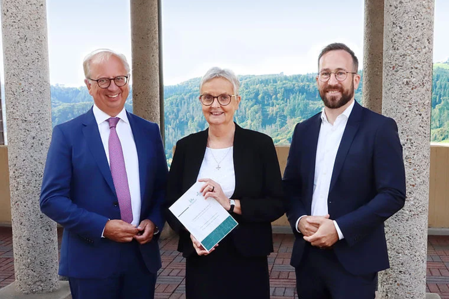 Drei Personen in formeller Kleidung stehen vor Säulen mit Waldlandschaft im Hintergrund, die mittlere Person hält ein Dokument
