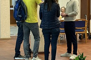 Vier Personen stehen in einem Raum mit braunem Boden, eine Person hält ein Mikrofon an einer langen Stange über eine andere Person, die spricht.