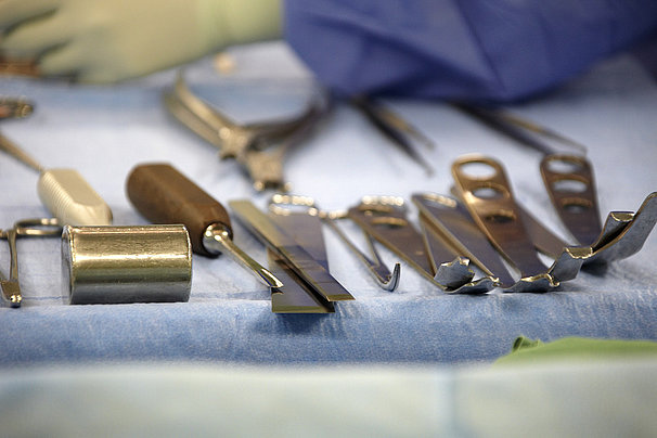Chirurgische Instrumente wie Skalpelle, Haken und Scheren auf einem blauen Tuch, im Hintergrund eine Hand mit Handschuh