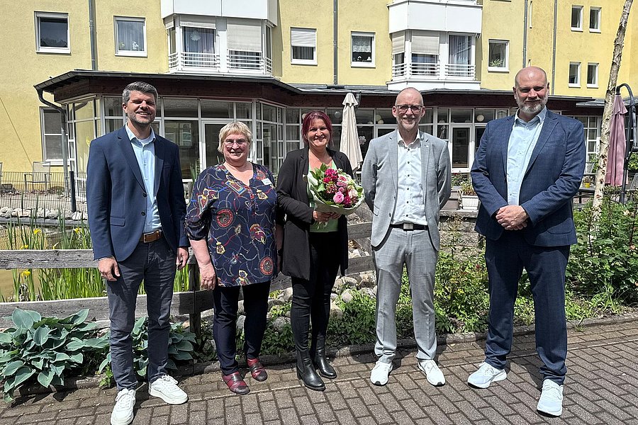 Fünf Personen posieren im Freien, umgeben von Pflanzen und einem Gebäude, einige halten Blumen.