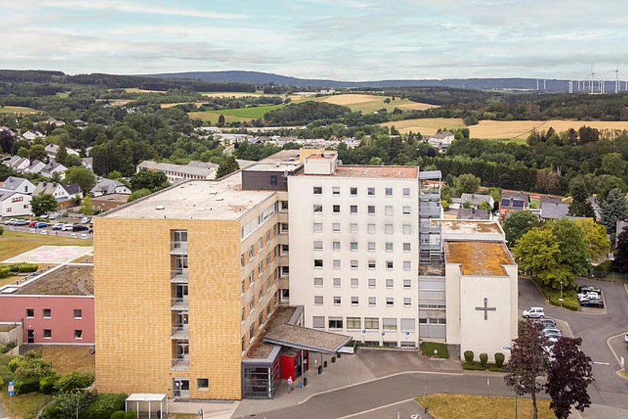 Krankenhausgebäude mit Kreuz auf der Fassade, umgeben von Bäumen und Wohnhäusern in einer ländlichen Gegend